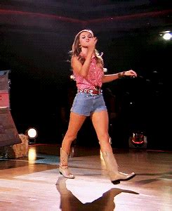 A Woman In Short Shorts And Cowboy Boots On Stage With Her Hand Up To