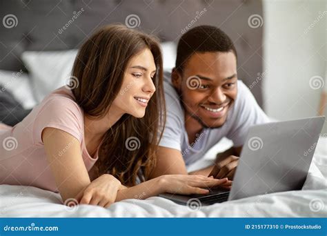 Interracial Loving Couple Spending Weekend Together At Home Using Laptop While Lying On Bed