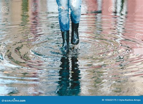 Womans Feet With Black Rubber Boots And Blue Jeans Standing In A Puddle Of Water After Rain On A