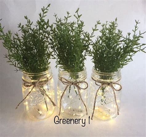 Three Mason Jars With Lights And Greenery In Them