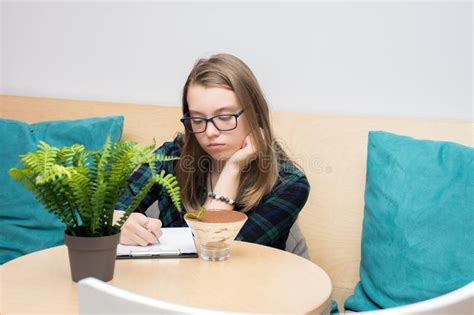 Pensive Teenage Girl In A Plaid Shirt And Glasses Writes In A Notebook