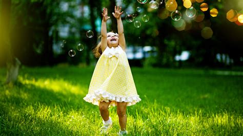 Cute Little Girl Is Playing With Bubbles On Green Grass Wearing Yellow