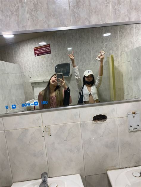 two women taking selfies in a bathroom mirror with urinals on the wall