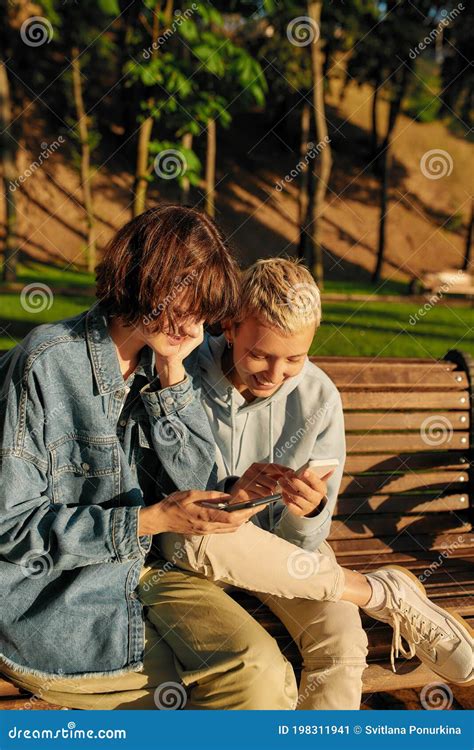 Two Joyful Women Holding Their Smartphone Sitting On The Bench In The Park Lesbian Couple