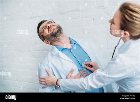 Female Doctor Listening To Heartbeat Of Colleague Who Has Heart Attack