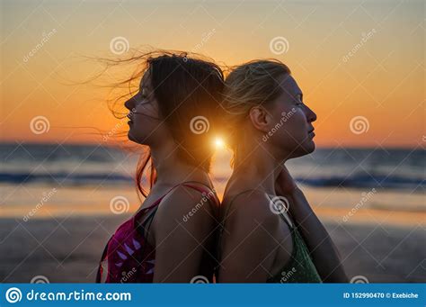 blonde and brunette girlfriends are clinging back to each other against sunset over the sea