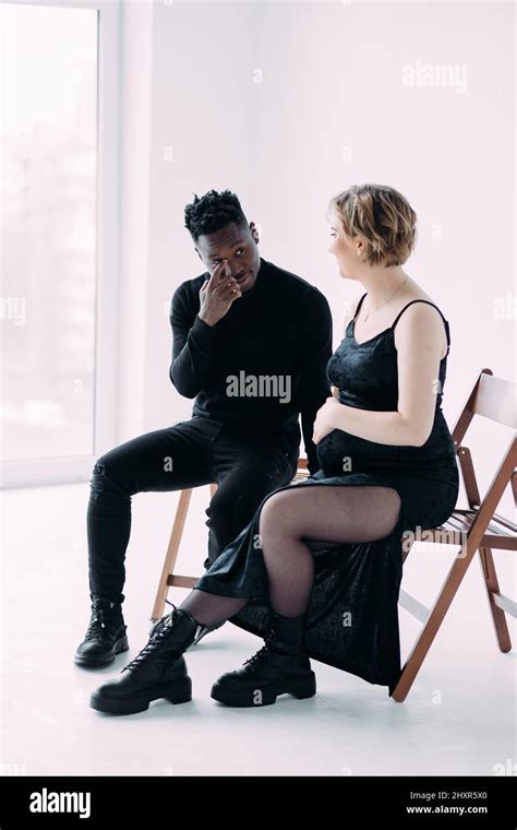 Young African Man And His Caucasian Pregnant Woman Sit On Chairs And Hold Hands With Each Other