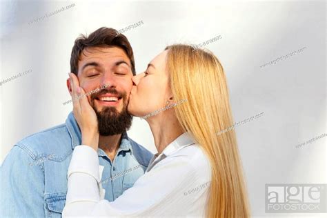 Affectionate Blond Woman Kissing Man On Cheek In Front Of Wall Stock