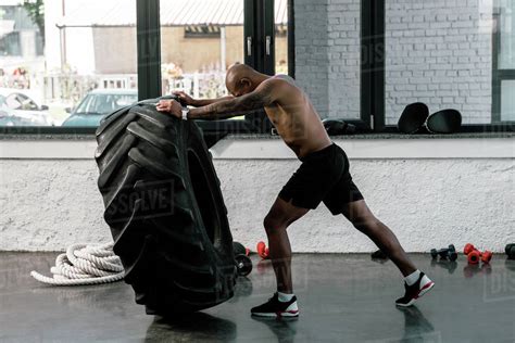 side view of muscular shirtless african american sportsman training with tire in gym stock