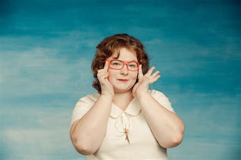 Attractive Red Haired Plump Woman In A White Suit Adjusts Red Glasses On A Blue Background Stock
