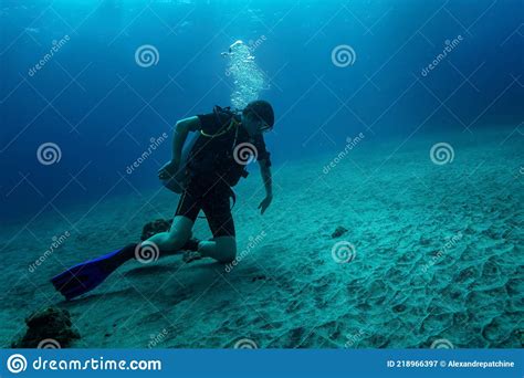 Young Diver Sits On Sandy Bottom On Knees And Carefully Looking At