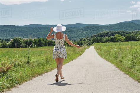 Back View Of Female Wearing Dress And Sunhat Walking With Bunch Of Flowers Along Road On