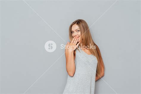 Portrait Of A Happy Young Woman Covering Her Mouth Isolated On A Gray