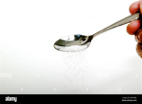 Grains Of White Sugar Pouring From A Silver Teaspoon Against A White