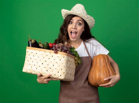Довольная молодая брюнетка женщина садовник в униформе в садовой шляпе держит корзину с овощами