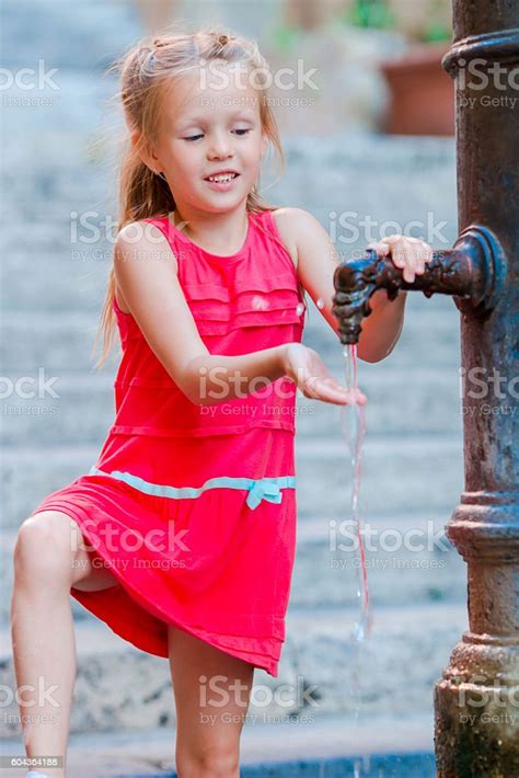 Little Adorable Girl Drinking Water From The Tap Outside At Stock Photo