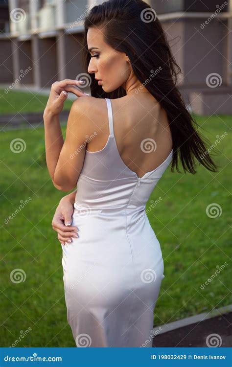 A Young Beautiful Brunette In A Light Summer Dress Poses On The Street With Her Back Turned