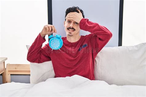 Hispanic Man Holding Alarm Clock In The Bed Stressed And Frustrated With Hand On Head Surprised