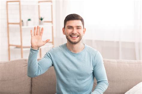 Millennial Guy Waving Hello Smiling To Camera Sitting On Sofa Stock
