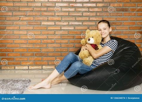 Beautiful Caucasian Teen Girl Sitting In Black Bean Bag Chair Holding Big Brown Teddy Bear Toy
