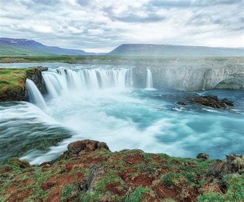 The Waterfall Is Surrounded By Green Mossy Rocks And Blue Water With