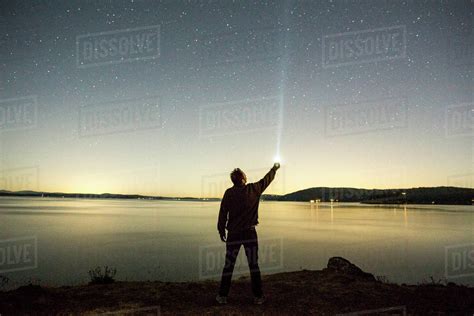 Caucasian Man Pointing Flashlight At Night Sky Near Water Stock Photo