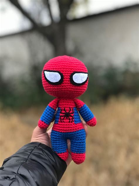 A Hand Holding A Crocheted Spider Man Doll In Front Of A Tree