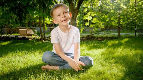 Cute Smiling Boy Sitting On Grass In Backyard Garden And Smiling In