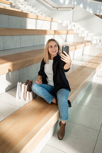 Stylish Blonde Girl Sitting In A Mall With Purchases Using A Mobile