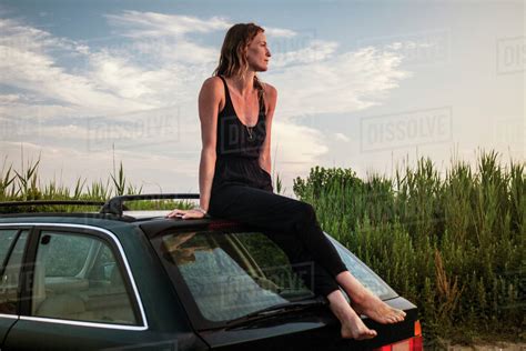 Thoughtful Woman Sitting On Car Roof By Plants Against Sky Stock
