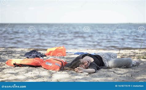 Body Of Dead Woman Lying On Seashore Near Life Jackets Shipwreck