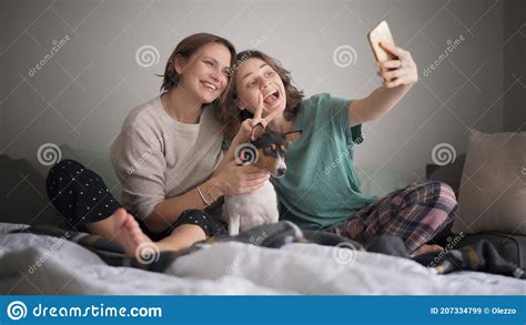 Beautiful Happy Lesbian Couple Sitting In Pajamas On The Bed In The Morning Making Selfie On