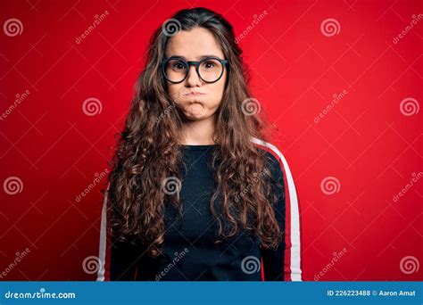 Young Beautiful Woman With Curly Hair Wearing Sweater And Glasses Over Red Background Puffing