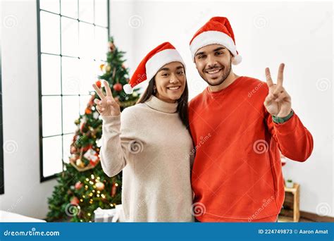 Young Hispanic Couple Standing By Christmas Tree Smiling Looking To The