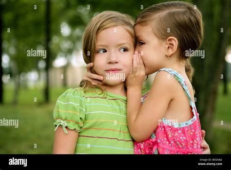 Two Twin Little Sister Girls Whisper In The Ear Outdoor On Green Park