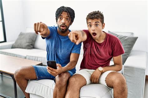 Young Hispanic Men Using Smartphone Sitting On The Sofa At Home Pointing With Finger Surprised