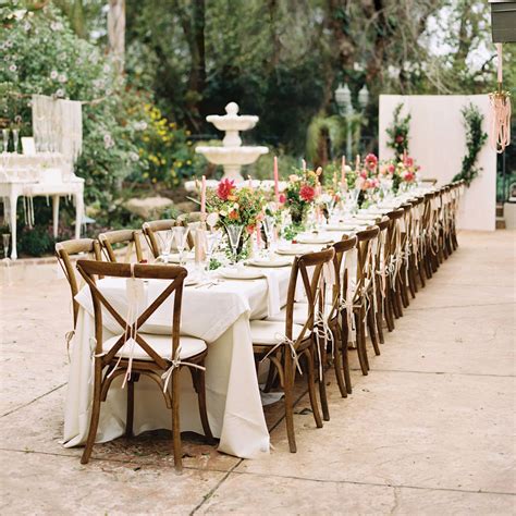 A Long Table Is Set Up With White Linens And Wooden Chairs For An Outdoor Dinner