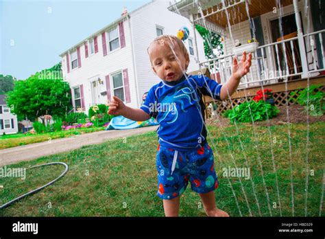 Caucasian Baby Boy Plays With A Water Sprinkler In His Front Lawn
