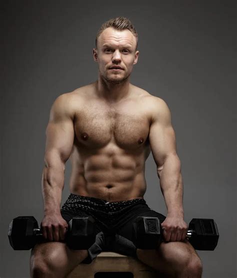 Handsome Power Athletic Man Training Pumping Up Muscles With Dumbbells In A Gym Barbells Behind