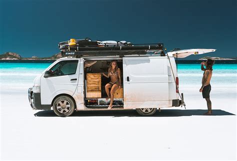 A Woman Standing Next To A Van With Surfboards On The Roof And Door Open