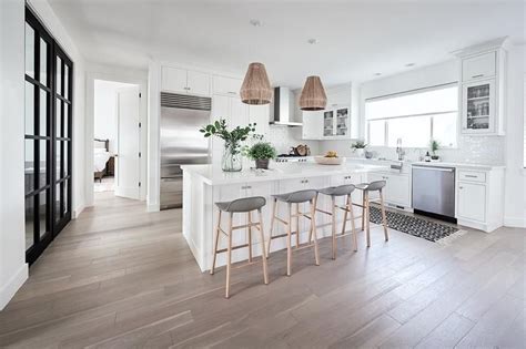 White kitchen interior with a wooden floor, a white bar stand, a row of white and gray stools, a laptop and consoles on. Low gray back stools sit at a white shaker kitchen island ...
