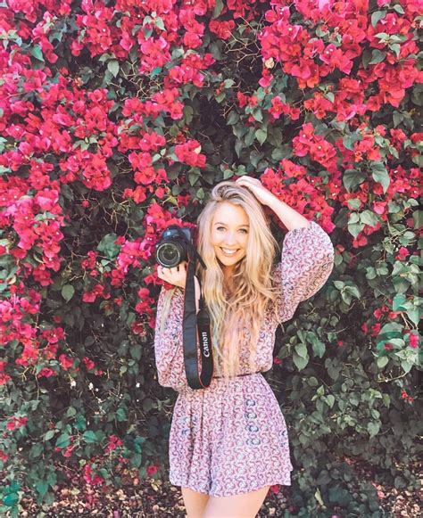 A Woman Standing In Front Of A Bush With Flowers Holding A Camera Up To