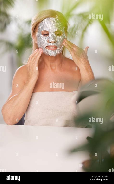 a senior woman putting beauty face mask on her face while sitting at home wrapped in towel