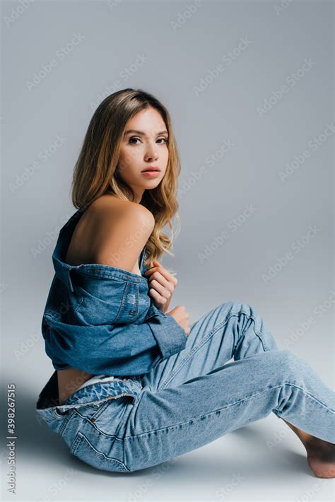 Sexy Young Woman In Unbuttoned Denim Shirt And Jeans Posing With Naked Shoulder On Grey Stock