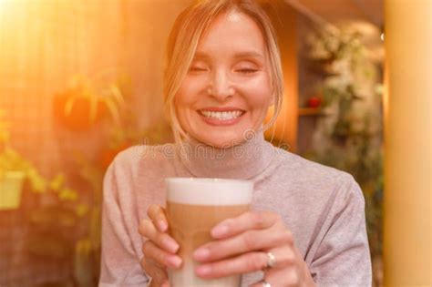Woman With Blonde Hair Sips Cappuccino In A Cafe She Is Holding The