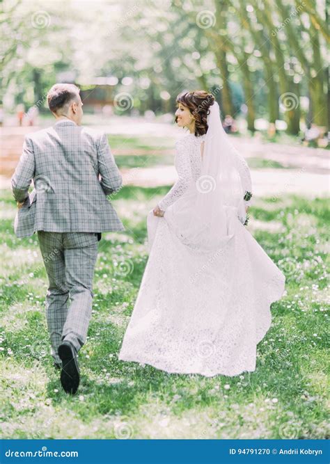 The Back View Of The Happy Newlywed Couple Running Along The Sunny Park