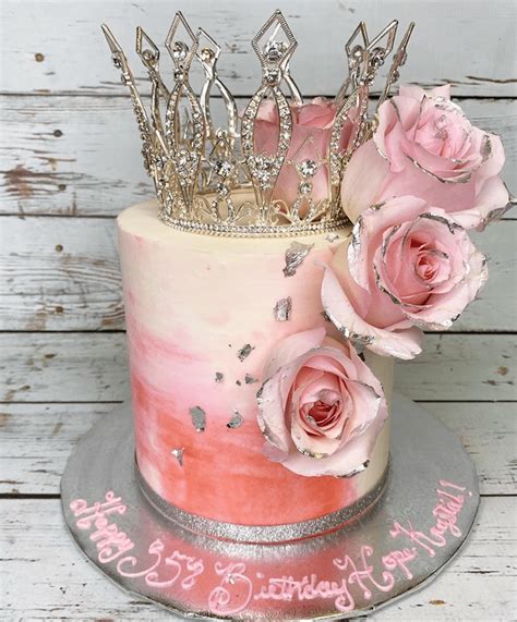 A Cake With Pink Frosting And Roses On The Top Is Decorated With A Crown