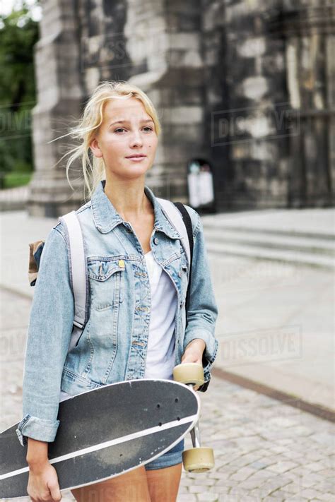 Teenage Girl Holding Skateboard Walking On Cobbled Street In City