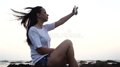 Beautiful Girl Takes A Selfie On A Smart Phone Sitting On The Beach Against The Evening Sky