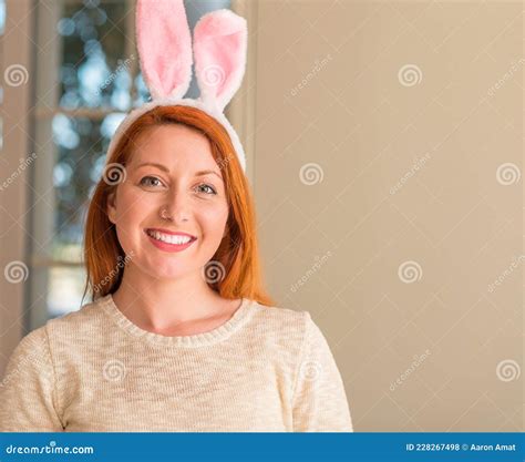 redhead woman wearing easter rabbit ears at home with a happy face standing and smiling with a